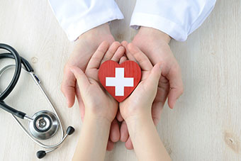 A child's hands in a professional's hands; the child is holding a red wooden heart with the medical symbol; a stethoscope at the side