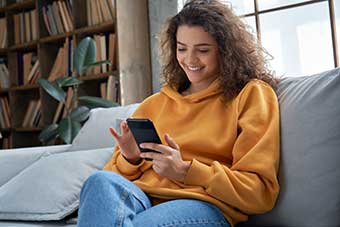 Happy millennial hispanic teen girl checking social media holding smartphone at home. Smiling young latin woman using mobile phone, relaxed on sofa.