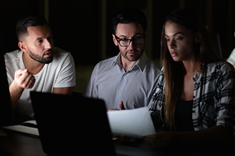 Group of young professionals collaborating in a dimly lit room, focused on a laptop screen, with others discussing and working in the background.