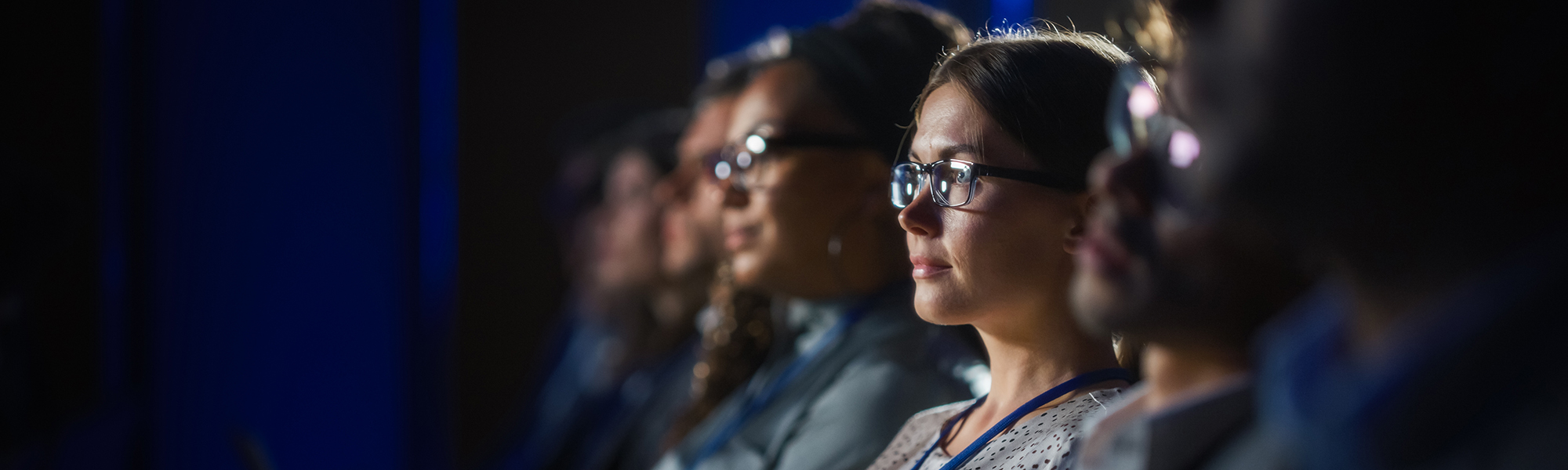 Audience members attentively watching a presentation or speaker in a dimly lit conference or seminar room, with a focus on a woman wearing glasses in the foreground.