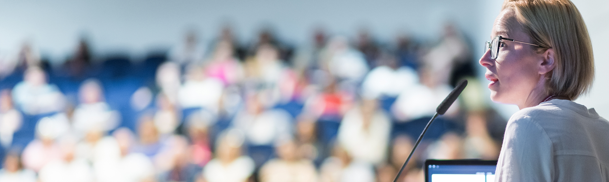 A woman with short blonde hair and glasses speaks into a microphone at a podium, addressing a blurred audience seated in the background.