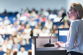 A woman with short blonde hair and glasses speaks into a microphone at a podium, addressing a blurred audience seated in the background.